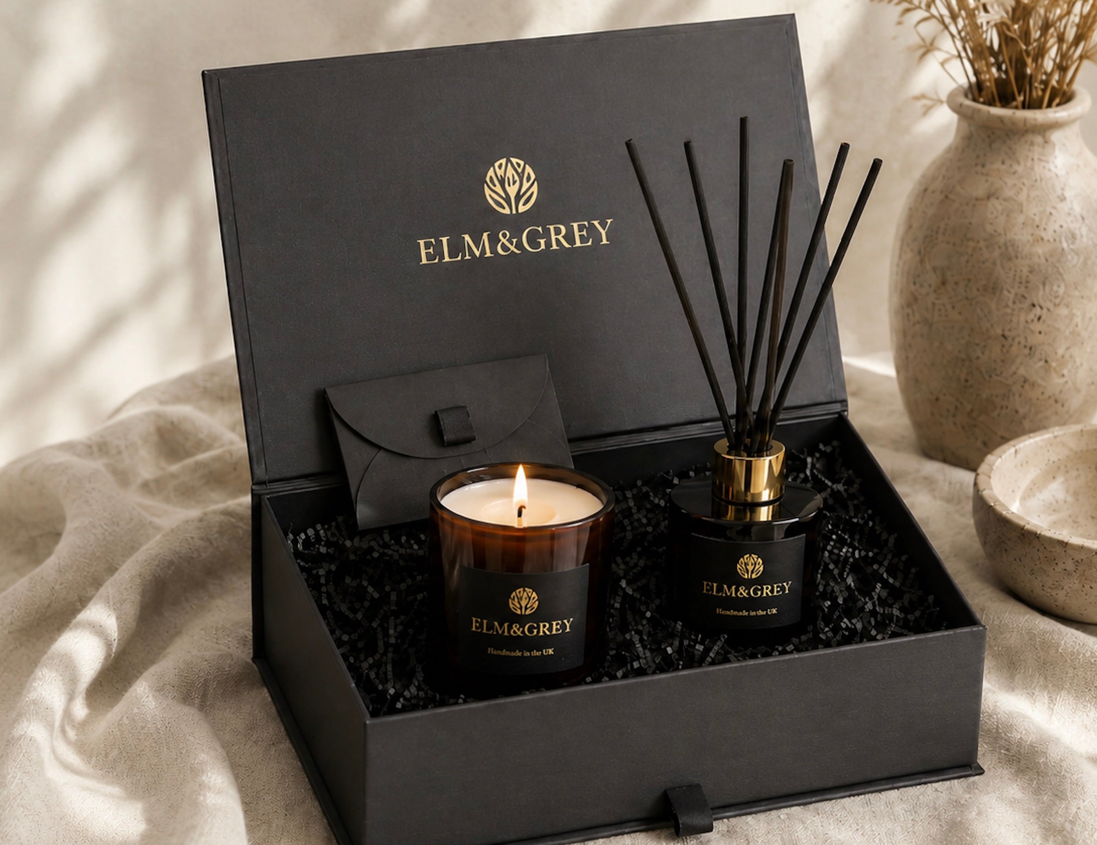 Open Elm & Grey gift box with candle and reed diffuser, styled on neutral linen in soft natural light, with black packaging and gold branding.