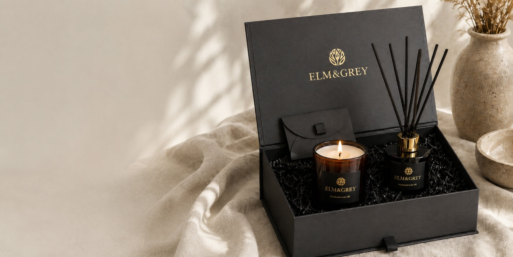 Open Elm & Grey gift box with candle and reed diffuser, styled on neutral linen in soft natural light, with black packaging and gold branding.
