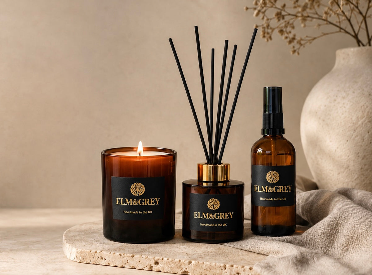 A set of ELM&GREY home fragrance products including an amber glass candle with a lit flame, a reed diffuser bottle with black reeds, and a spray bottle, all arranged on a stone slab with a neutral background.