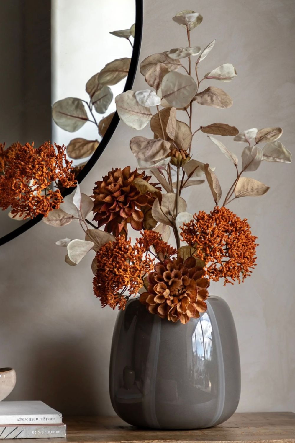 Grey vase with autumnal dried flowers and leaves on a wooden surface against a neutral wall.
