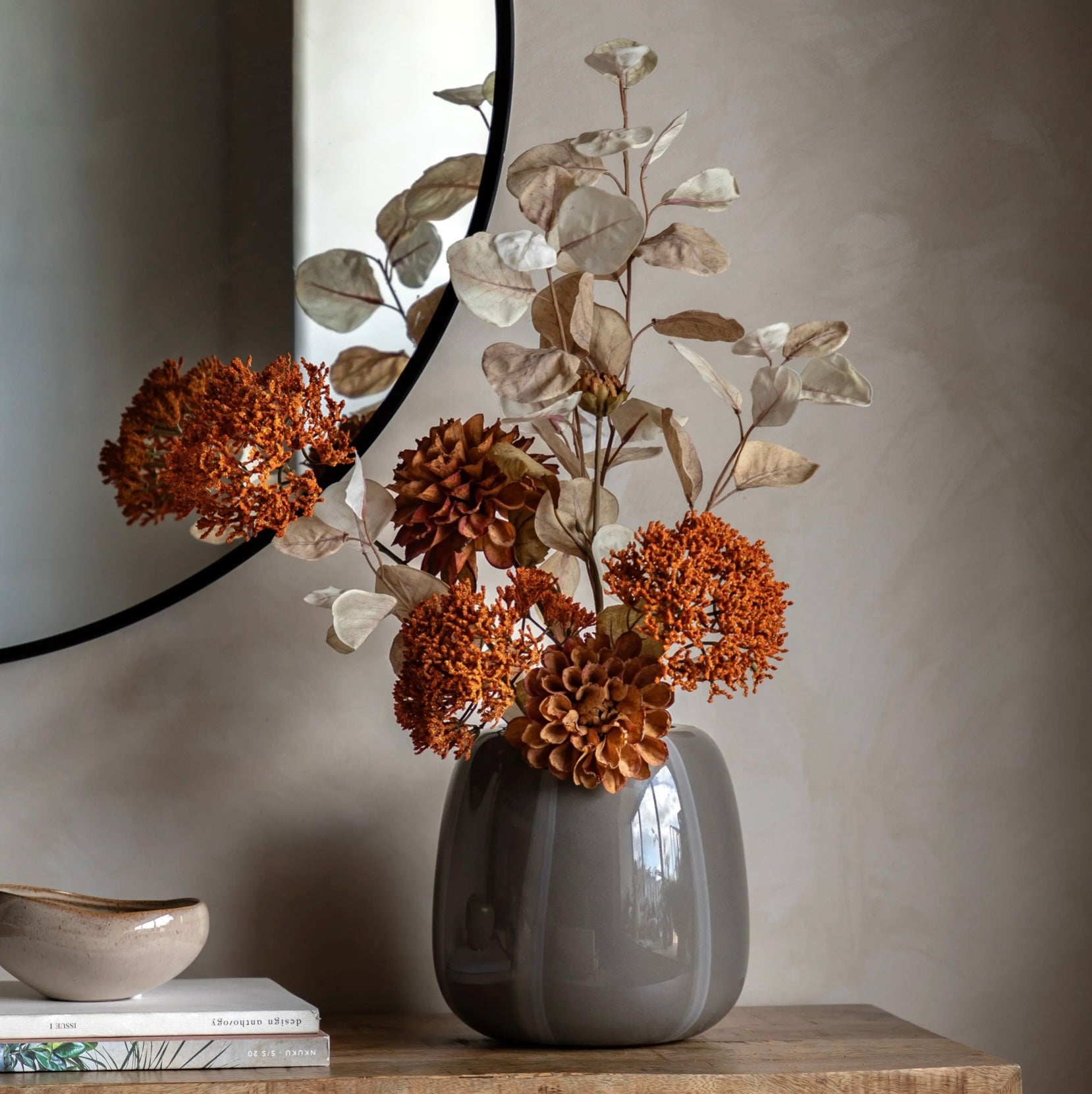 Grey vase with autumnal dried flowers and leaves on a wooden surface against a neutral wall.