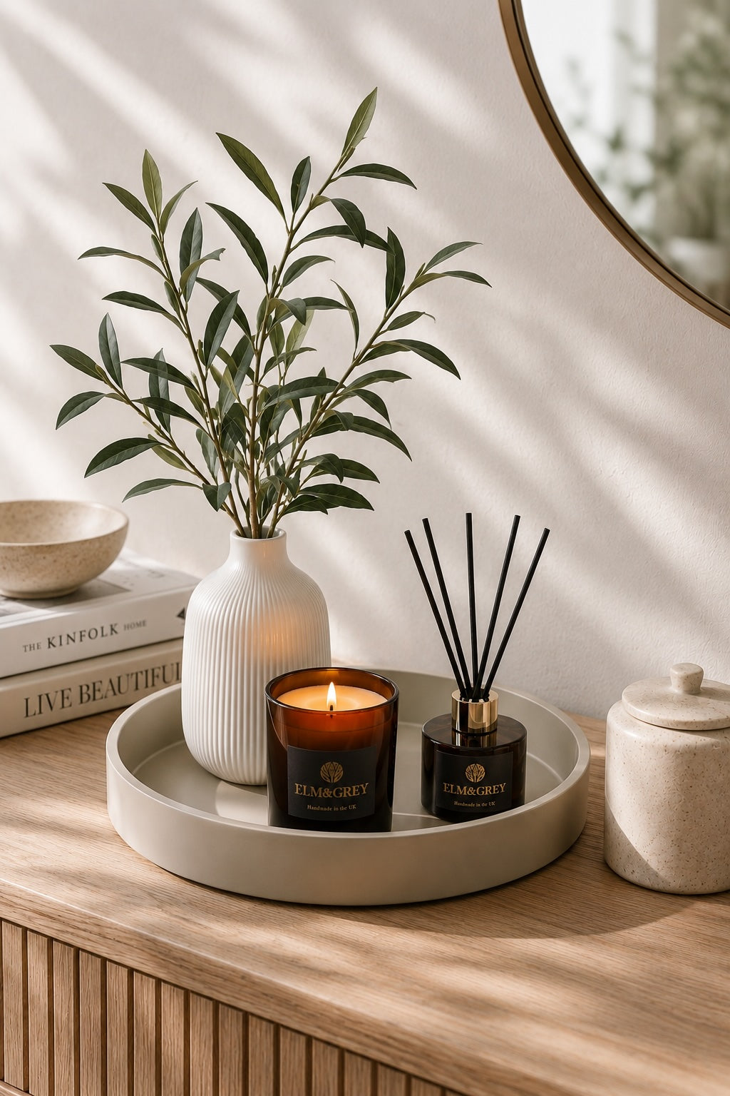 Faux olive arrangement in a white textured vase styled on a fawn round tray with luxury amber candle and reed diffuser, creating a calm and elegant home styling display