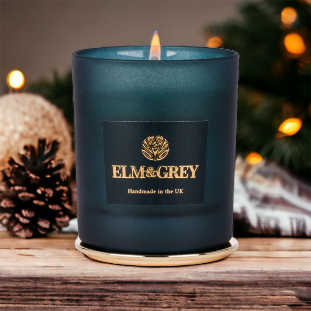 A blue glass candle vessel with a lit candle, branded ELM&GREY, on a wooden surface with holiday decorations in the background.