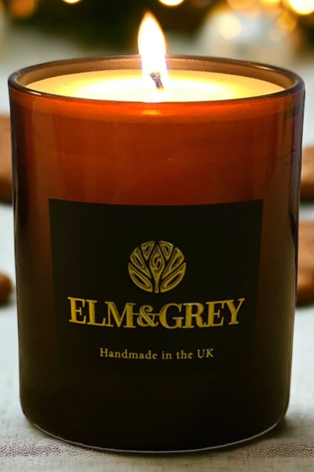 A brown scented candle with a lit flame, placed on a table with cookies and a decorative gingerbread man in the foreground.