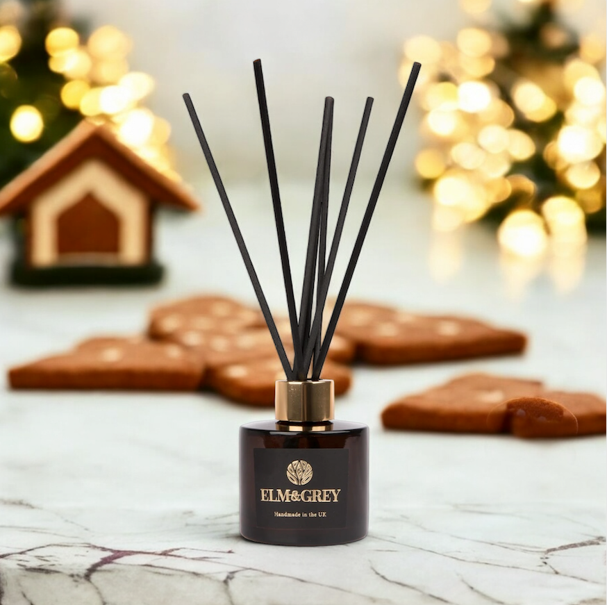 A black reed diffuser with a label reading 'ELM & GREY' on a white surface, surrounded by cookies and a festive decorated background.
