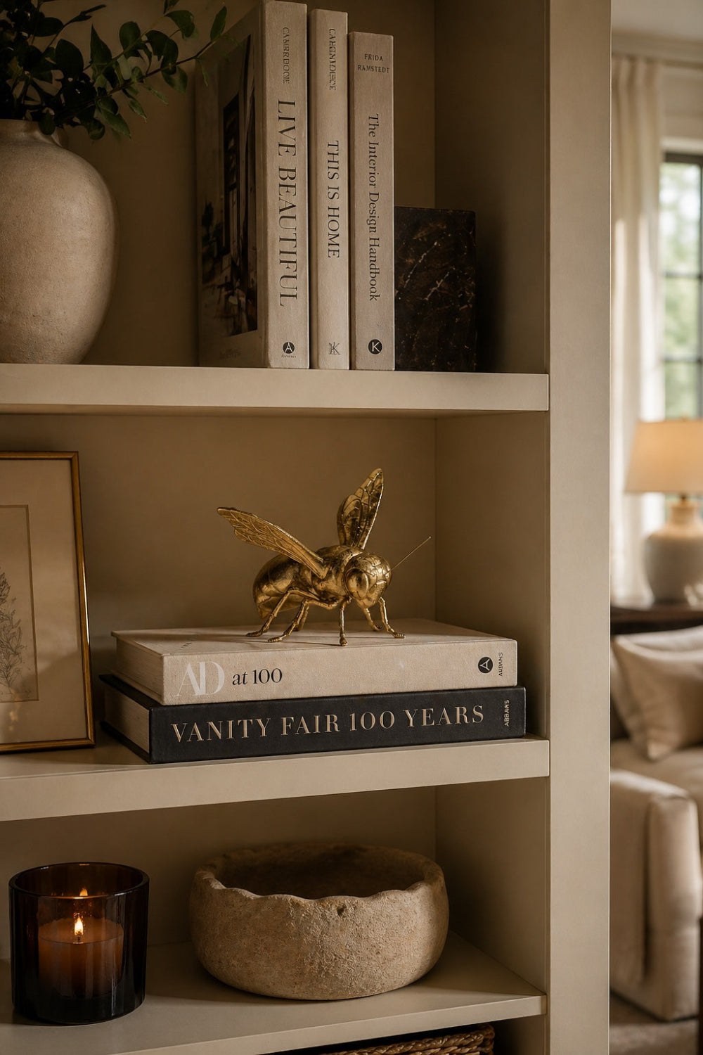 Gold decorative bee sculpture on stacked AD at 100 and Vanity Fair books on a styled bookshelf with lit candle and stone bowl — Elm & Grey