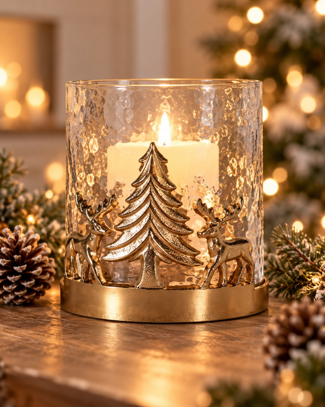 A festive holiday candle holder with a hammered clear glass cylinder, featuring a gold-toned metal base with a Christmas tree and two deer silhouette design. A white pillar candle is lit inside, casting a warm glow.