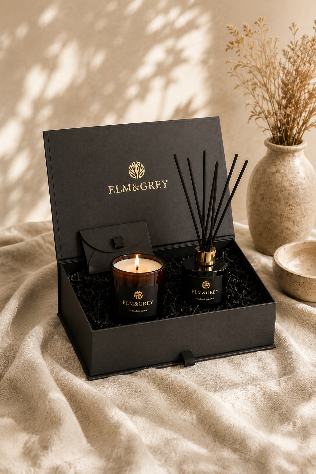 ELM&GREY candle and reed diffuser gift set in an open black box, styled on a beige textured blanket with a vase of dried stems in the background.