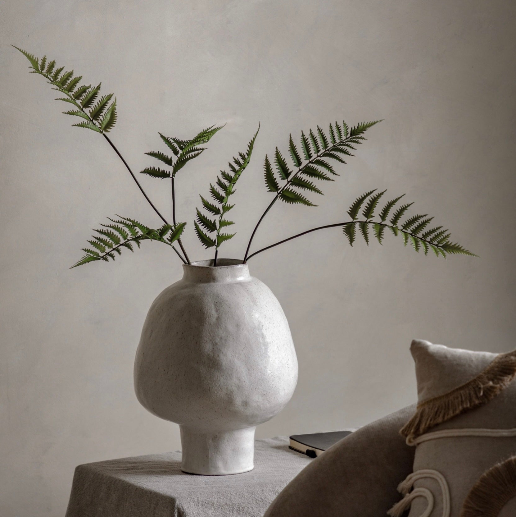 White textured vase with ferns on a table against a neutral wall