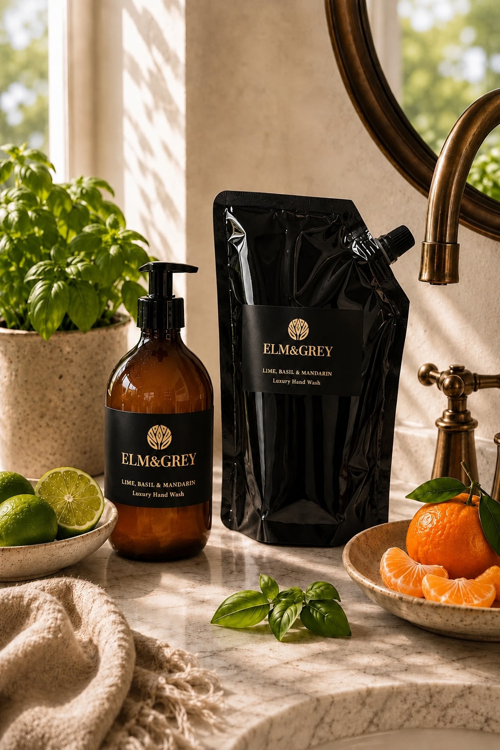 Elm & Grey Lime, Basil & Mandarin luxury hand wash in an amber glass pump bottle with matching refill pouch, styled on a marble countertop beside fresh limes, basil leaves and mandarin in a warm sunlit bathroom setting.