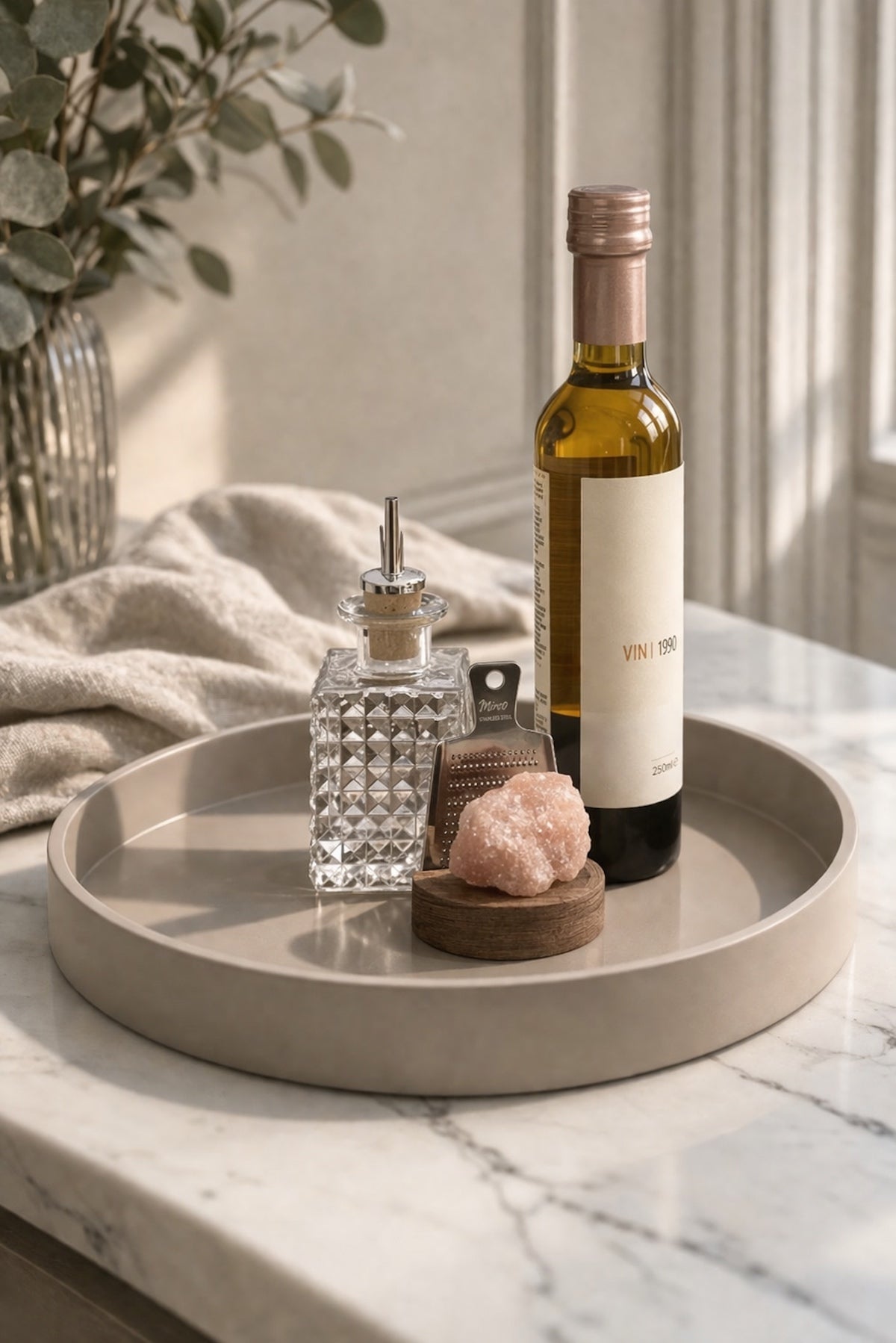 Elm and Grey image with a bottle of oil on a mojoo lacquered tray resting on a marble surface with decorative items and a plant.