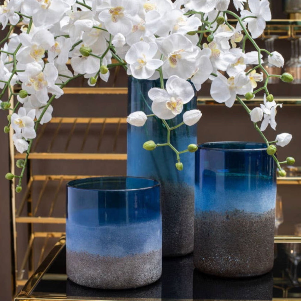 Blue sand effect gradient vases with white orchids on a reflective surface.
