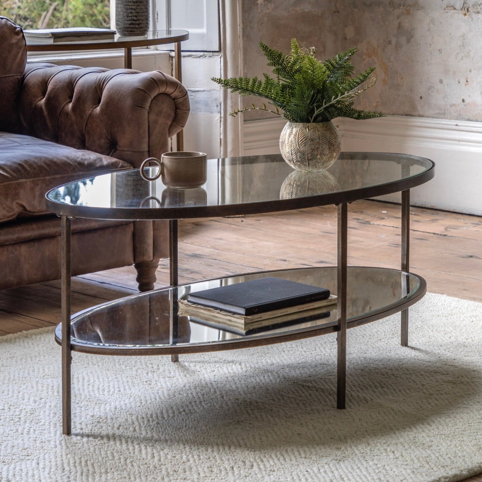 Glass and bronze oval coffee table with books and a plant in a living room setting