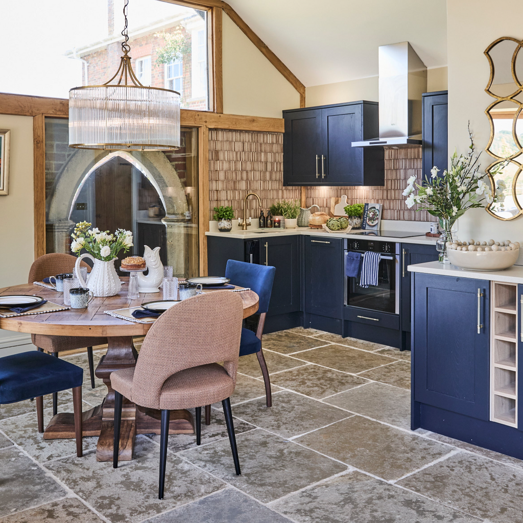 A kitchen with blue cabinets and chanderlier and table laid with a glass cake stand gluggle jug and  white urchin jug with a flower arrangement,  ACream bobble vase and artifical faux flowers arrangement in white
