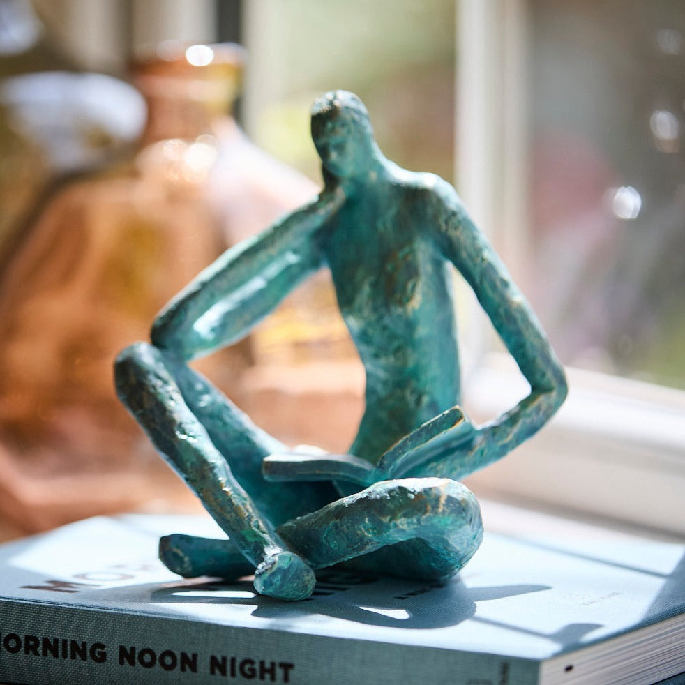 A verdigris decorative man sat reading a book by a window 