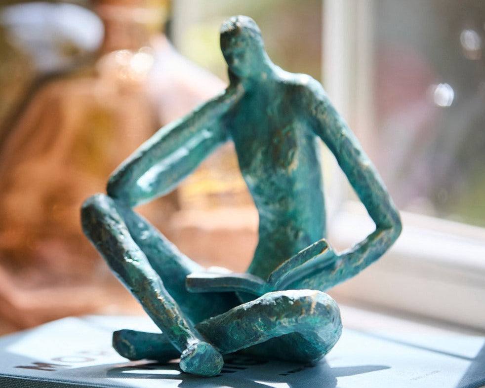 A verdigris decorative man sat reading a book by a window 