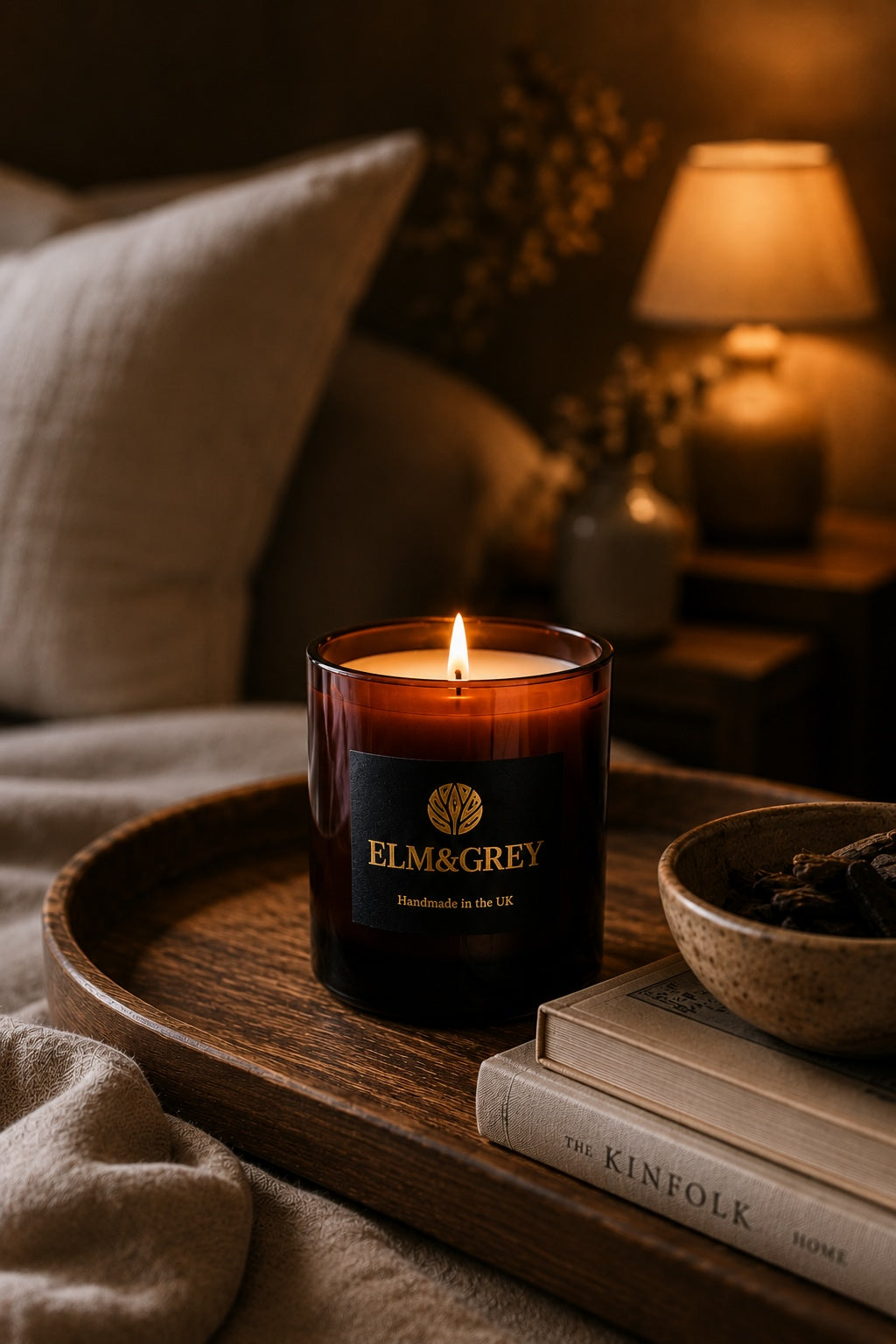 Amber lit candle in a glass vessel on a wooden tray with books and a bowl in a cozy room setting.