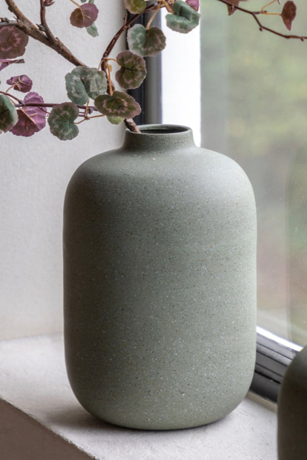 Olive green matte vase on a windowsill with a branch and leaves in the background