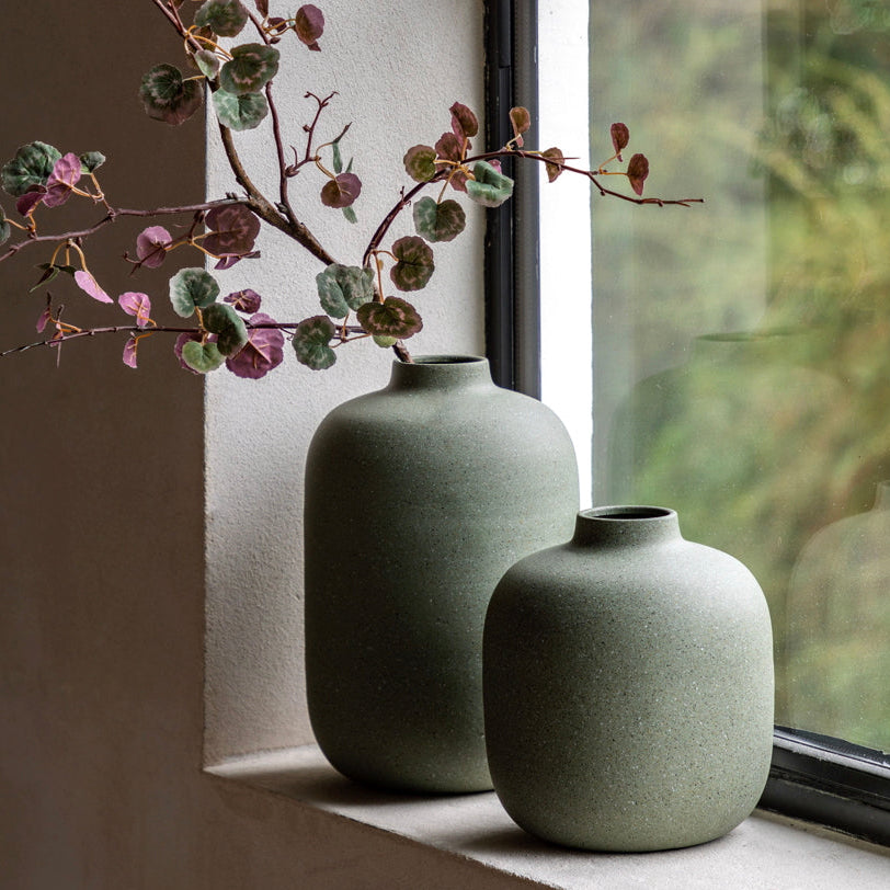 Two olive green vases on a windowsill with a branch of eucalyptus.