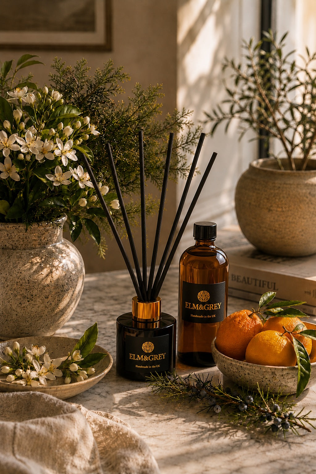 Orange Blossom & Juniper reed diffuser refill and reed diffuser by Elm & Grey, displayed on a marble surface with oranges, blossom flowers and warm sunlight, creating a fresh and uplifting home fragrance scene.