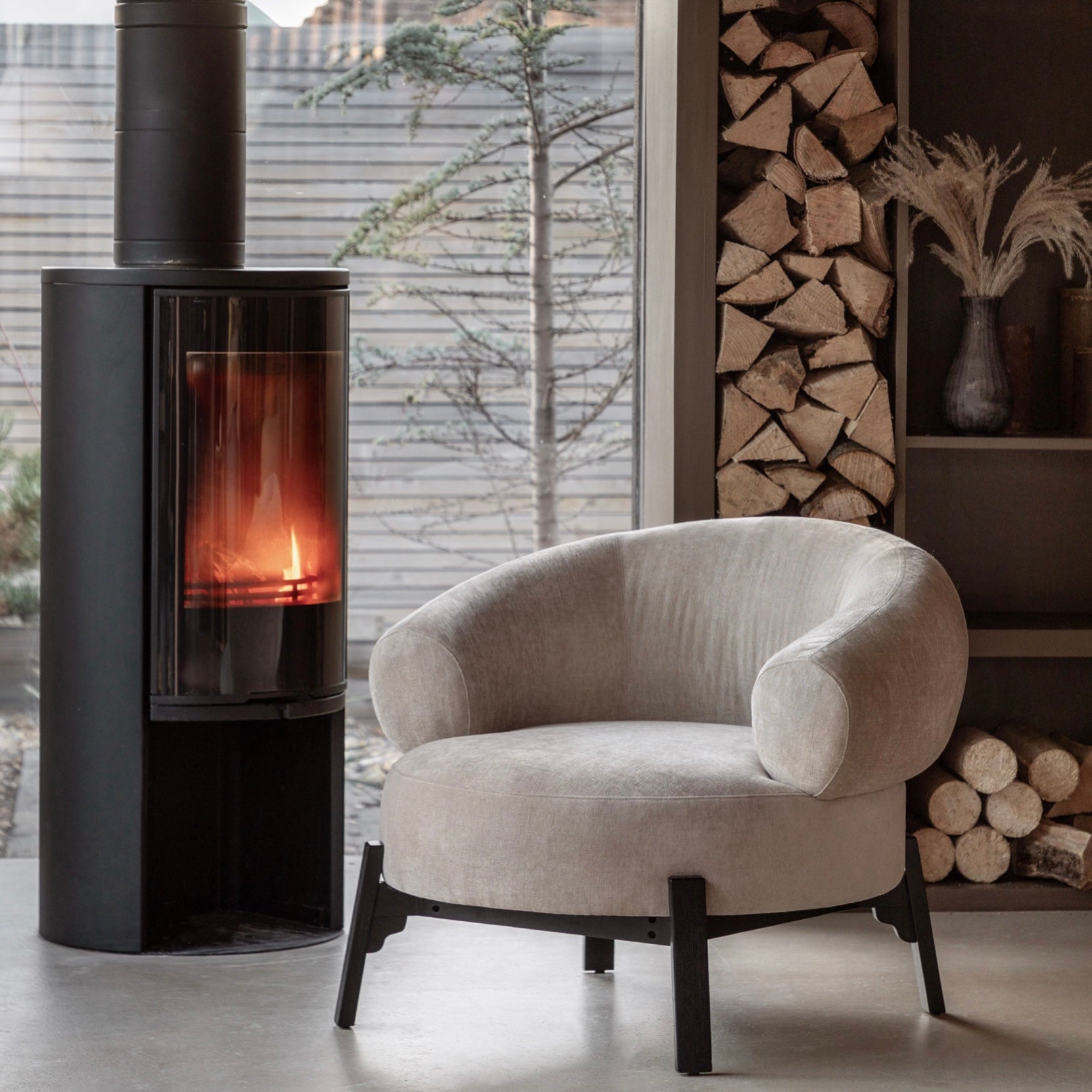 Modern curved armchair in a room with a stove and stacked firewood, Elm & Grey.