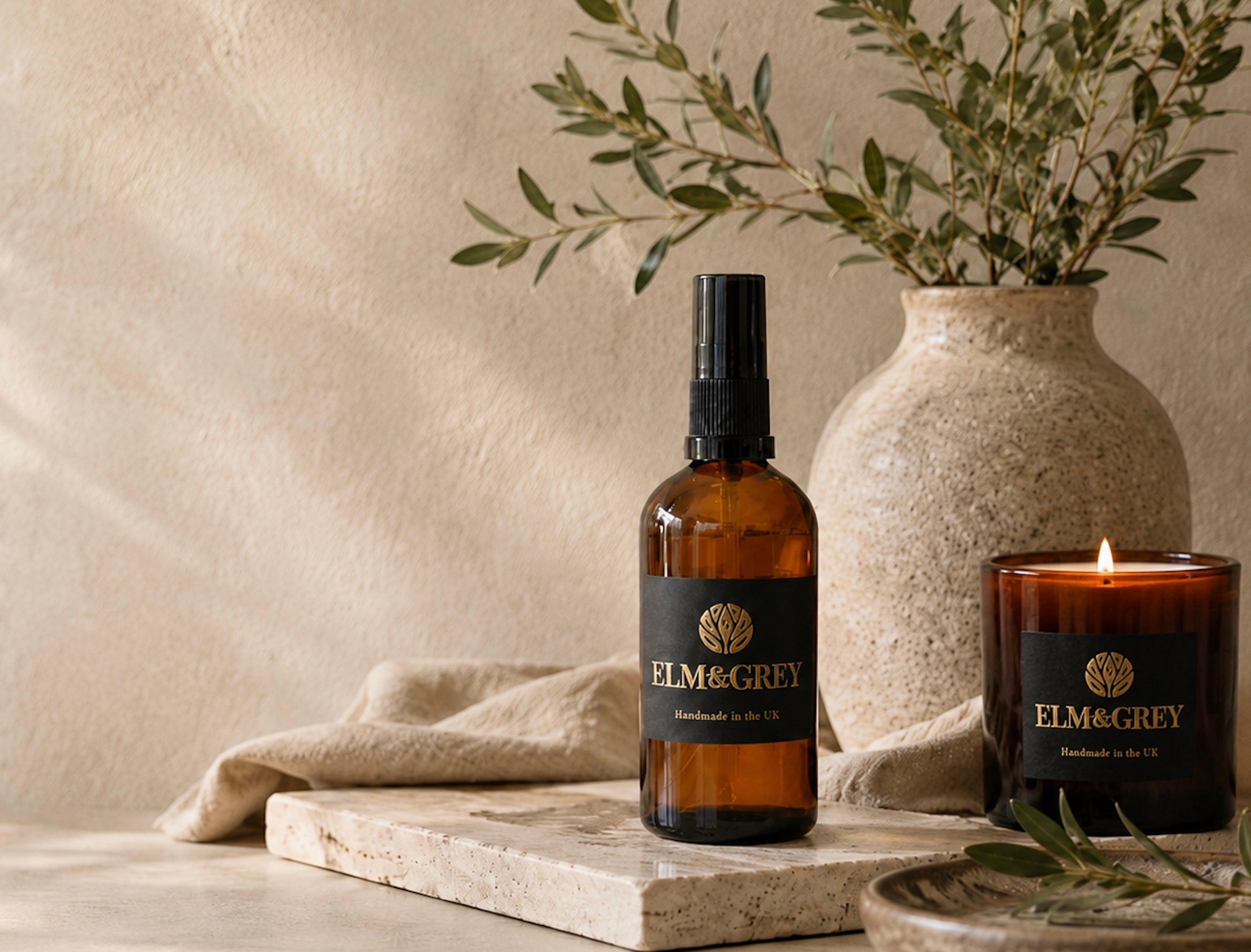 elm and grey room spray and lit candle in amber vessels in front of a stoneware vase and olive branches resting on a marble slab