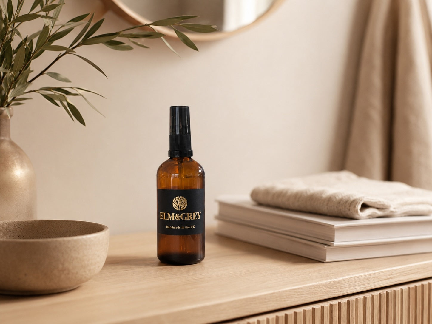 elm and grey lifestyle image ofa room spray on a console table alongside neutral coloured books, bowl and olive branches