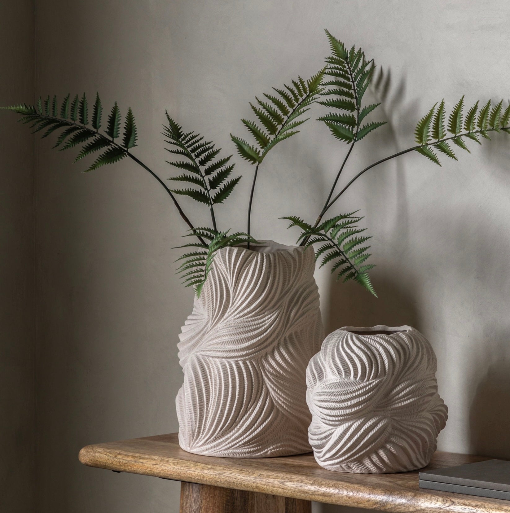 Two textured ceramic vases with ferns on a wooden surface against a plain wall.