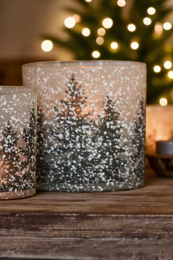 Two candle holders with tree silhouettes on a wooden surface with blurred lights in the background