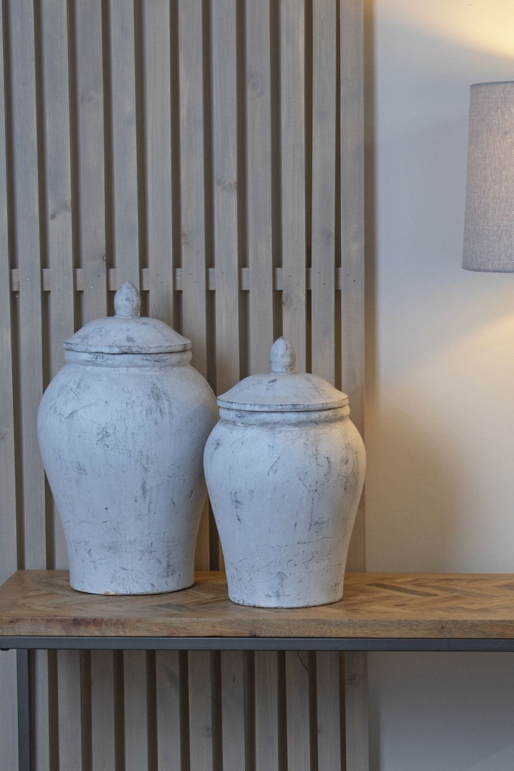 Two white ceramic jars with lids on a wooden shelf against a slatted wall.