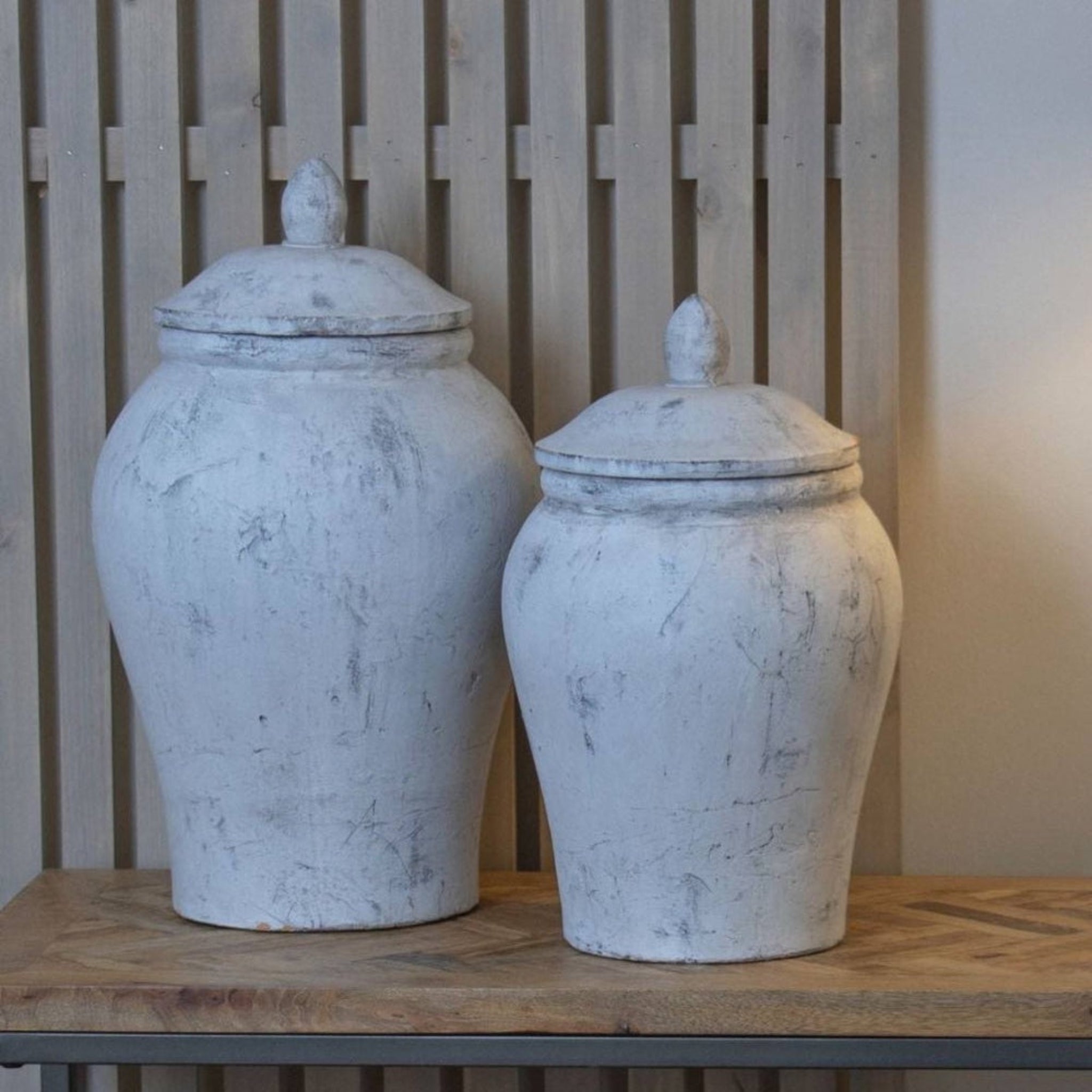 Two white ceramic jars with lids on a wooden surface against a wooden panel background