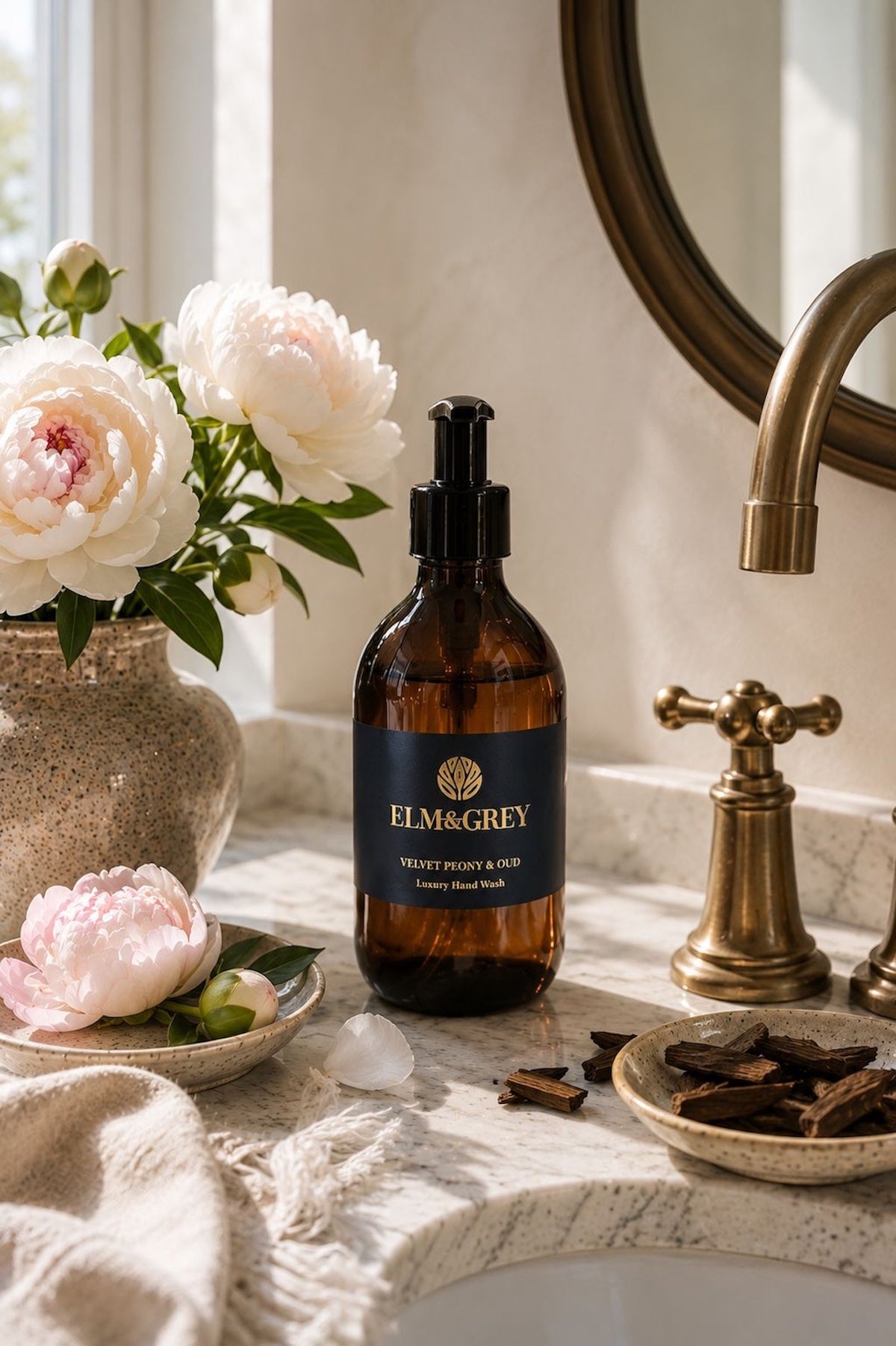 Elm & Grey Velvet Peony and Oud luxury hand wash on marble bathroom surface with blush pink peonies and oud wood chips beside brass tap