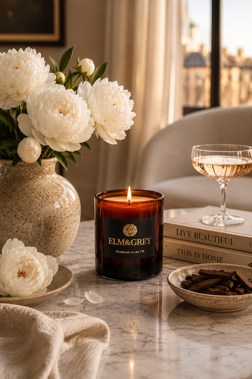 Elm & Grey velvet peony and oud scented candle on a table with flowers, books, and a glass of champagne.