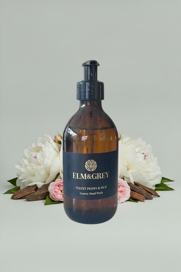 Amber glass bottle with pump dispenser labeled 'Elm & Grey' on a light grey background with flowers.