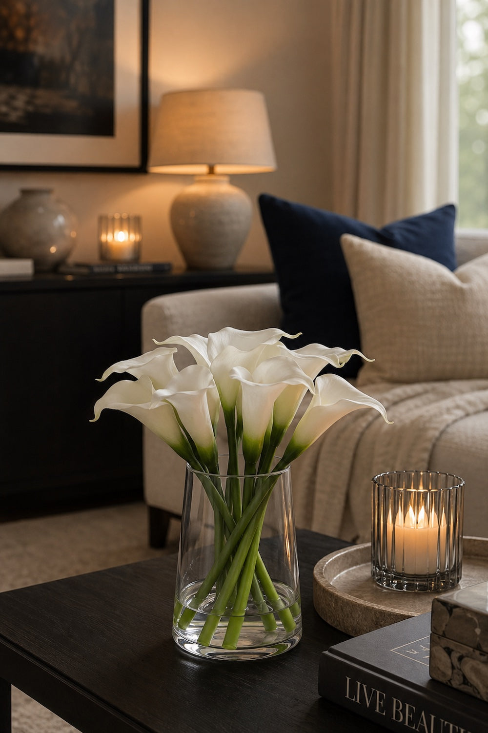 White calla lilies in a clear glass tapered vase on a dark wood coffee table with lit ribbed tealight and Live Beautiful book in an evening living room setting — Elm & Grey