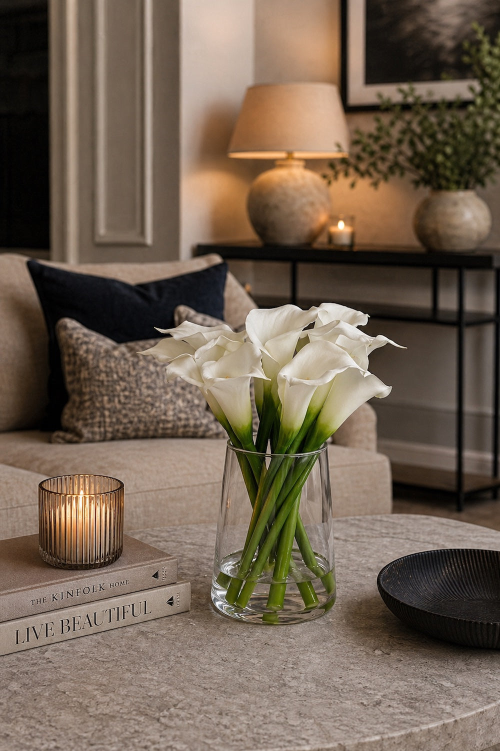 White calla lilies in a clear glass tapered vase on a travertine coffee table with Kinfolk books and lit ribbed tealight in a warm neutral living room — Elm & Grey