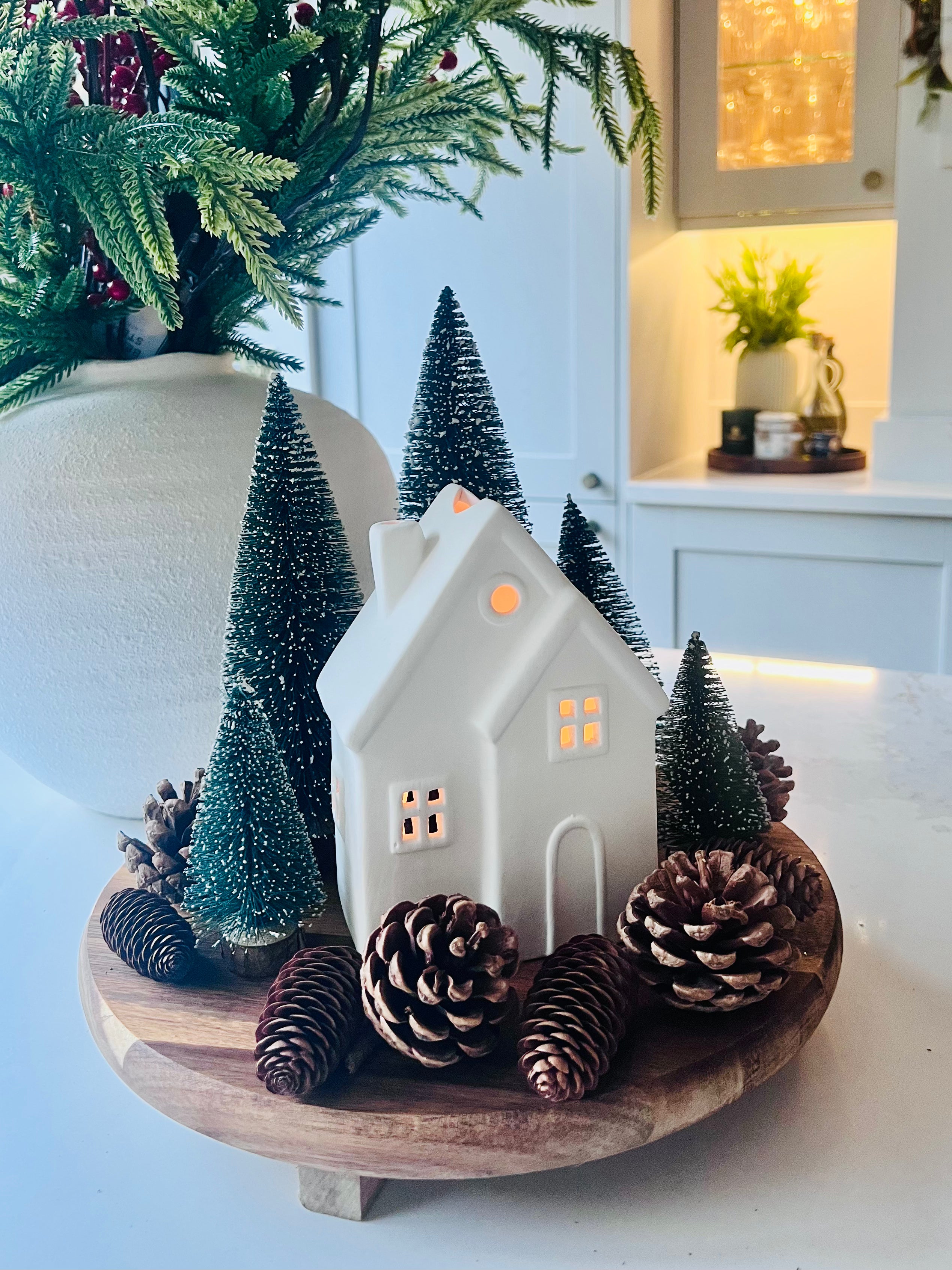 Decorative setup with a white ceramic house, pinecones, and small trees on a wooden stand.