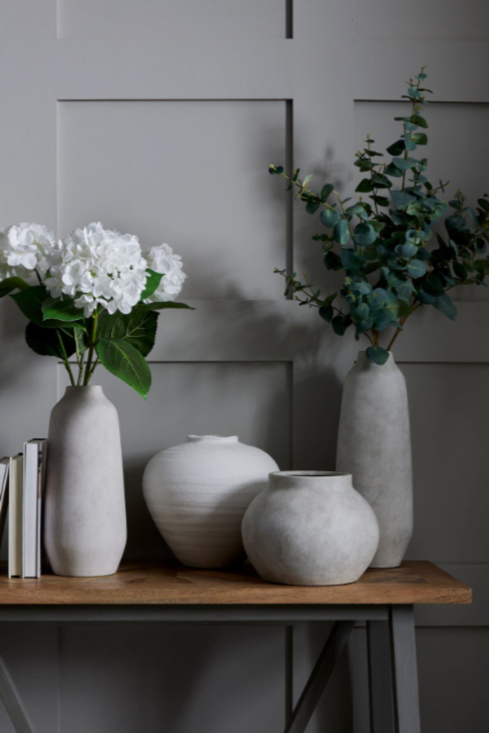 White ceramic stone vases and a plant on a wooden table against a gray wall.
