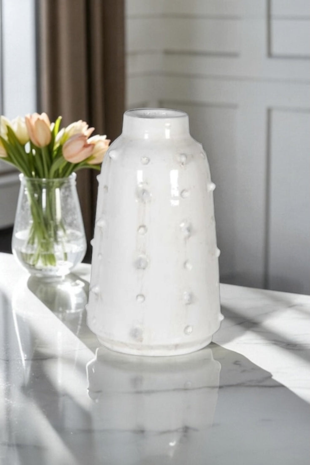 White textured vase on a marble surface with a smaller clear vase containing pink tulips in the background.